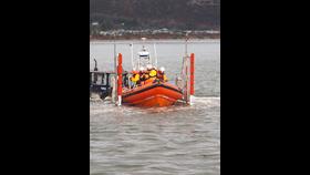A lifeboat carrying several crew members in yellow and red uniforms is being deployed into the water from a vehicle on a ramp. The scene takes place near the shore with hills and buildings visible in the background.