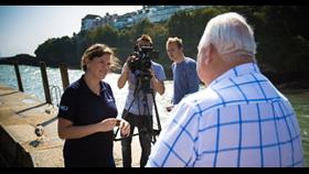 women interviewing man on harbour with camera man in background.