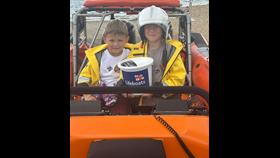 two young boys sat on an RNLI lifeboat with holding a collection bucket.