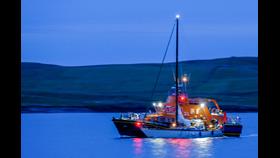 An image of Lerwick Lifeboat at sea coming into harbour at mightnight, with the yacht secured alongside on the port side, in the semi-darkness