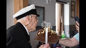 Ronald Quested in full Naval uniform blowing out candles on a chocolate cake with happy birthday topper held by his daughter as he celebrated his 98th birthday
