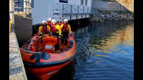 Four crew on board an inshore lifeboat tied up at a quay wall with the lifeboat station visible in the background. The crew are standing facing the camera with sunshine and blue sky.