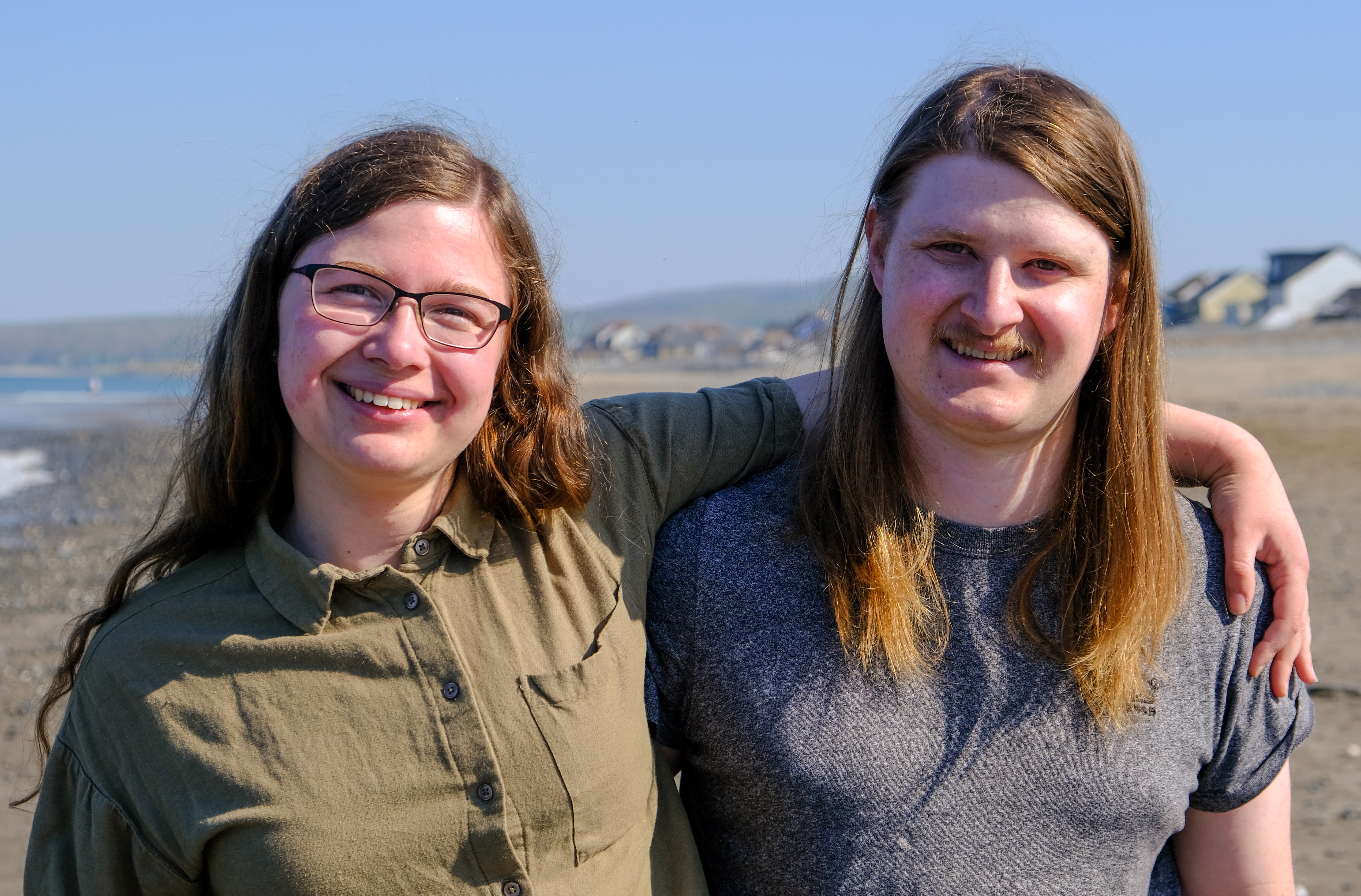 Katie and Will smiling on a beach with their arms round each other