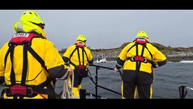 Three lifeboat crew members in full all-weather yellow suits and red lifejackets on the bow of the lifeboat
