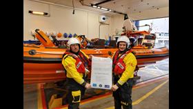 Two crew in drysuits and lifejackets in the boat house holding an open day poster while standing in front of the inshore lifeboat.