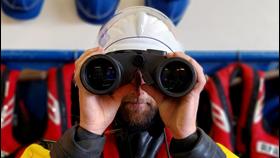 Close up of the head of a crew member in a drysuit and wearing a helmet and looking through a pair of binoculars towards the camera