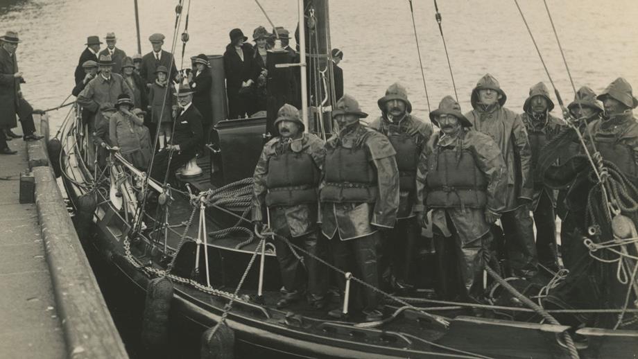 Black and white image of lifeboat crew in kapok lifejackets and kit onboard lifeboat with group of children and adults