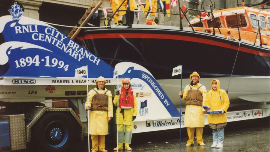 Colour image of four people dressed in historical crew kit with lifeboat behind, taking part in procession