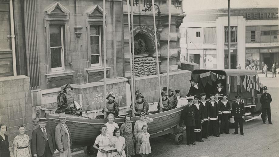 Black and white image of lifeboat pulled by van, lifeboat crew inside with oars raised and fundraisers alongside