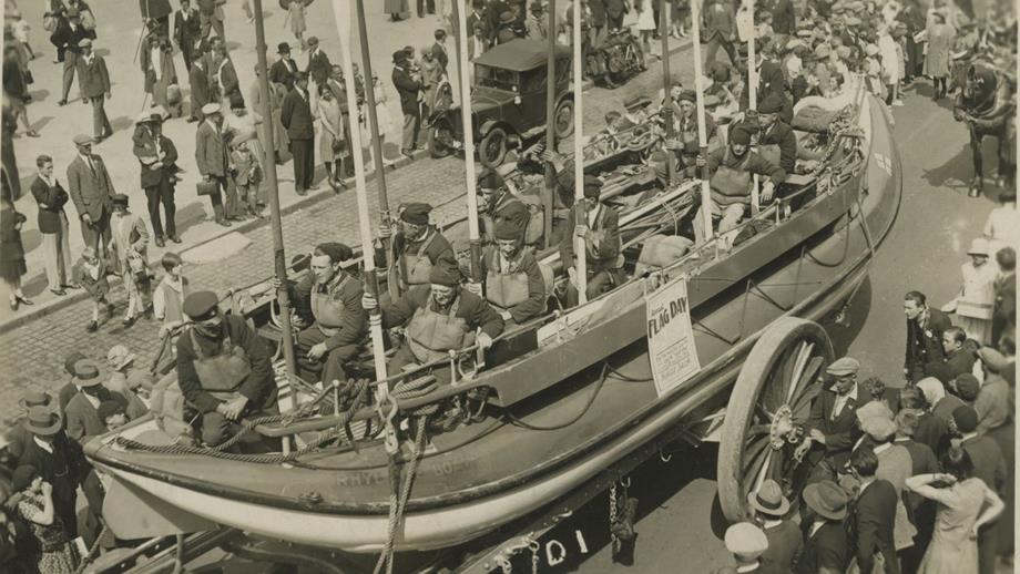 Black and white image of lifeboat being pulled through crowded street with crew inside