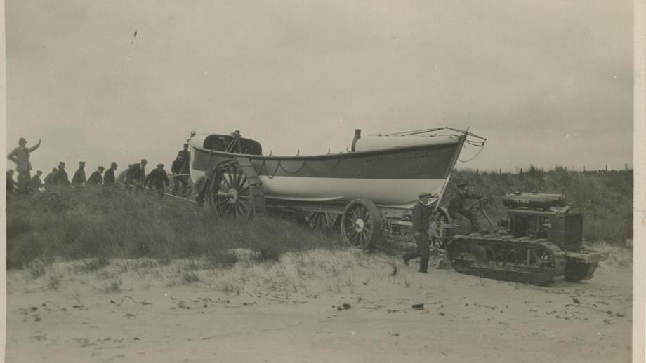 Black and white image of tractor towing lifeboat over sand dune