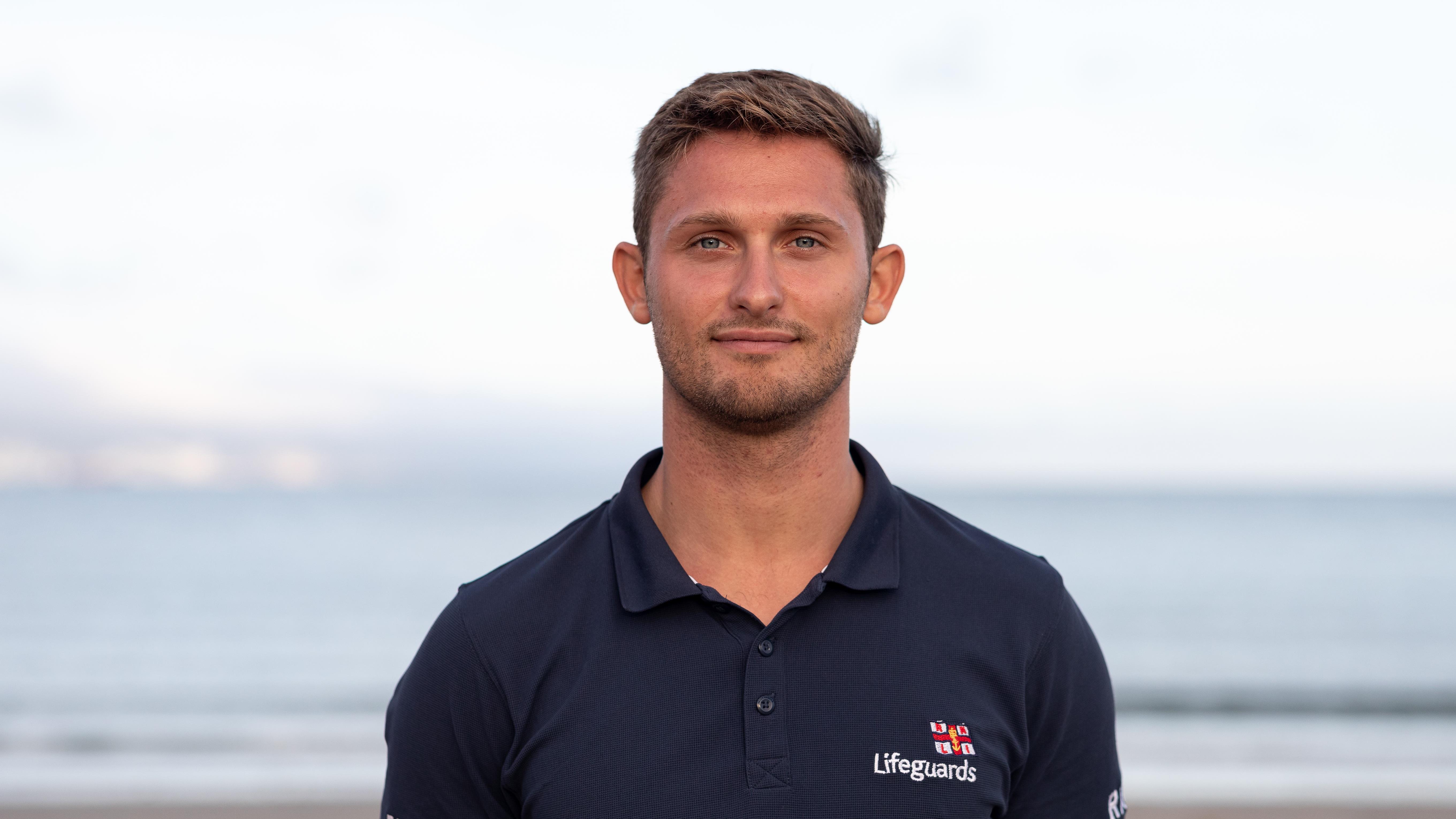 RNLI Lifeguard Supervisor Dom Fajkiel smiling, with the sea in the background.