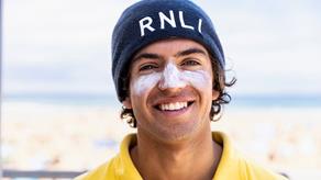 A head and shoulders shot of RNLI Lifeguard Harry Barea. Harry is smiling and wearing a blue RNLI woolly hat, a yellow RNLI polo short, and sunscreen across his nose and cheeks. The beach in the background is deliberately blurred.