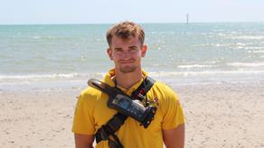 RNLI Senior Lifeguard Zeph Palmer in a lifeguard tshirt smiling at a sandy beach