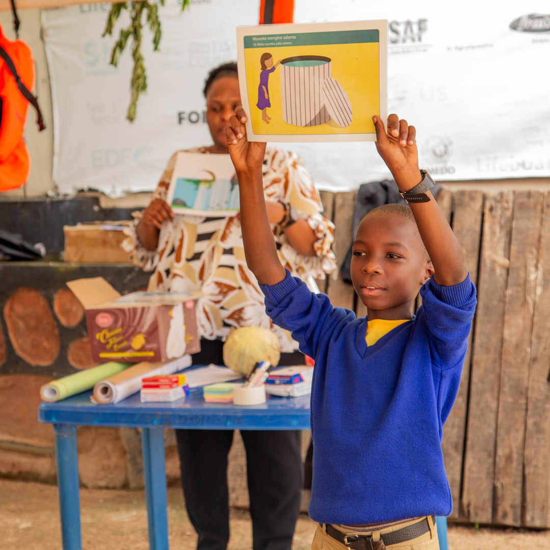 A schoolchild is standing up in front of his class, holding up a poster. He is wearing light brown trousers, and a bright blue jumper with a yellow T-shirt underneath. There is a teacher behind him, who is standing at a table with some art supplies such as pens, paper and tape. The teacher is also holding up a poster.