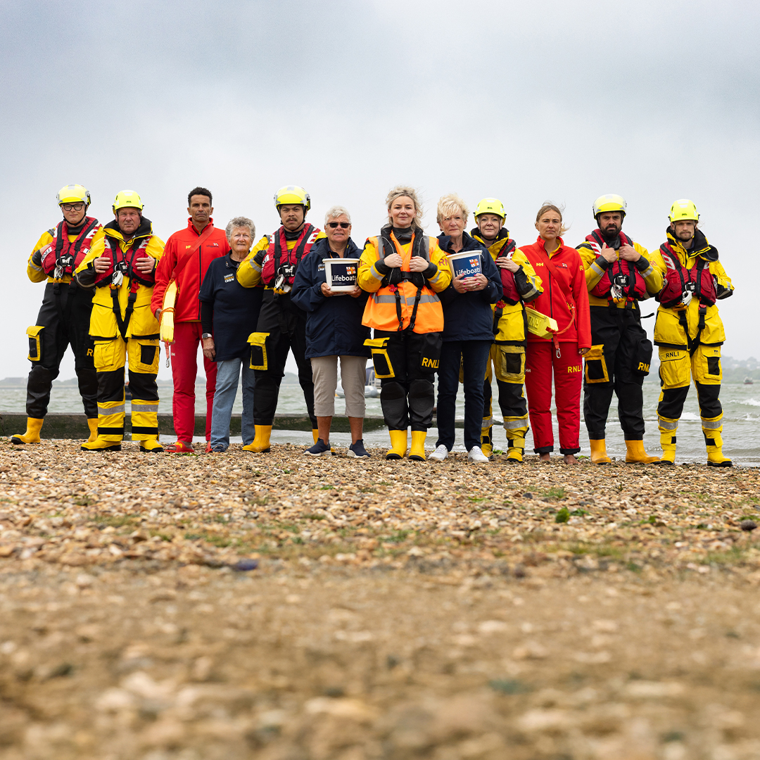 A group of RNLI lifesavers are standing in a line on a pebbly beach, all looking at the camera. Some of them are smiling, and some of them have a neutral expression. There are crew members, lifeguards and fundraising volunteers in the photo. The sea is behind them, and the sky is cloudy.