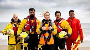 A group of five RNLI lifesavers are standing in a line, all looking at the camera. They all have a neutral expression on their faces. Some of them are wearing all-weather crew kit. One of them is wearing inshore crew kit. One of them is wearing a red RNLI lifeguard fleece. The sea is behind them, and the sky is cloudy.