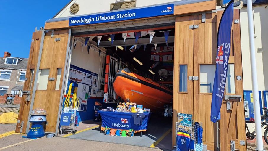 Orange lifeboat inside lifeboat station with "Newbiggin RNLI" sign, wooden doors, and RNLI merchandise for sale.