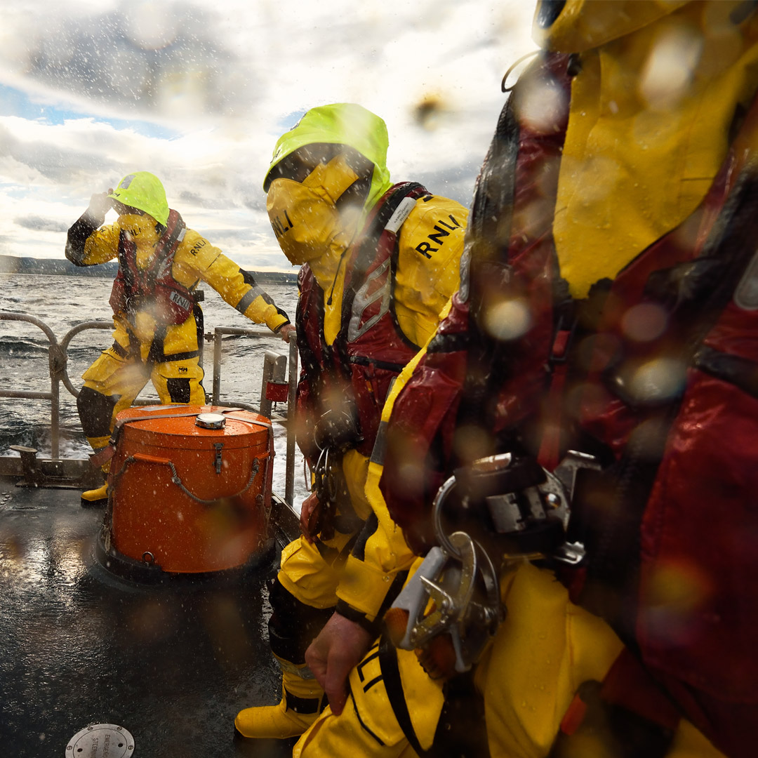 Three lifeboat crew members in full kit brace against rough seas and spray on the rear deck of an all-weather lifeboat