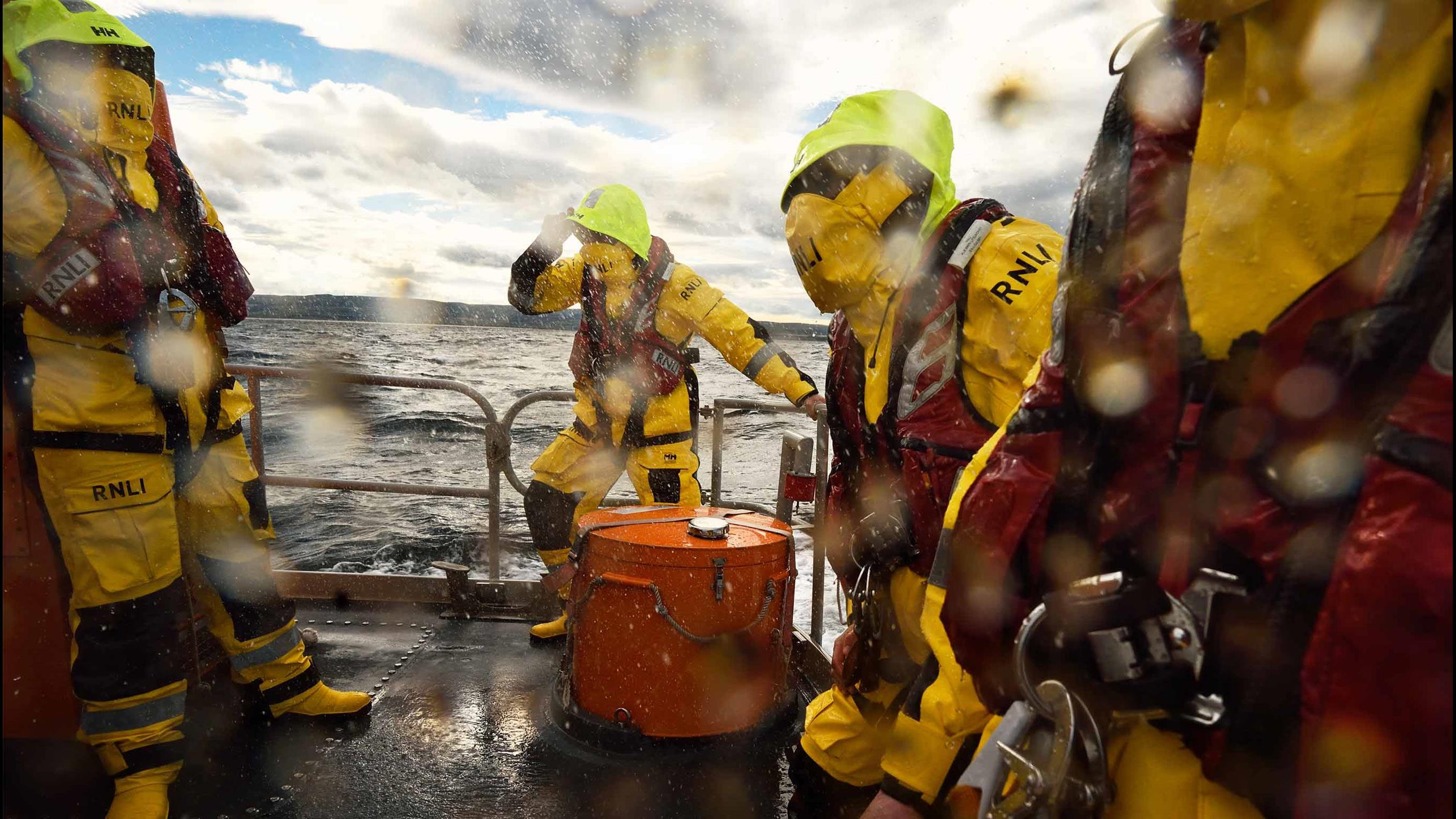 Four lifeboat crew members in full kit brace against rough seas and spray on the rear deck of an all-weather lifeboat