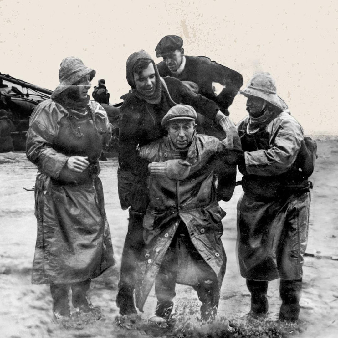 Black and white photo of lifeboat crew members carry injured men out of the water and onto the beach