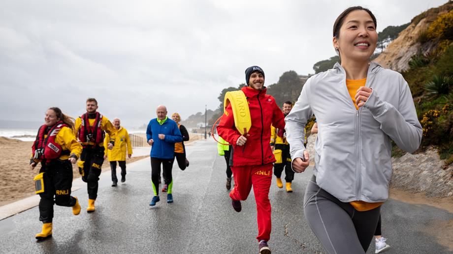 A group of people are running in a staggered pack along a beach promenade. Some of them are wearing running kit and others are wearing full RNLI lifeguard kit and full RNLI crew member kit