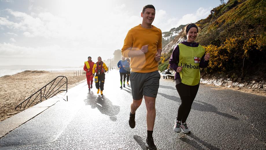 Two people are running side-by-side along a beach promenade. One is wearing an RNLI yellow vest