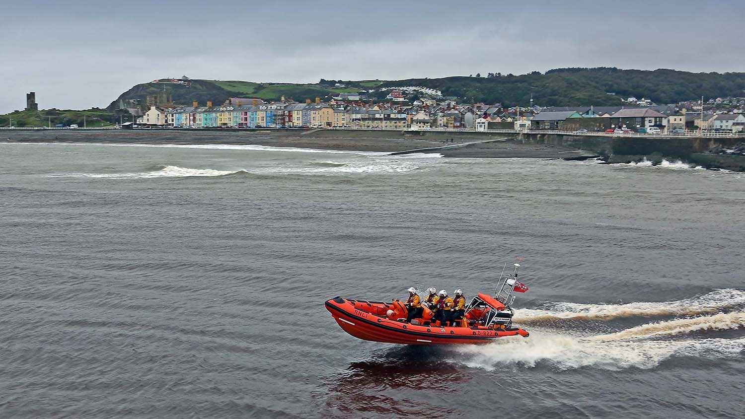 A B class lifeboat with four lifeboat crew members onboard is at sea on an overcast day, with houses and land in the background