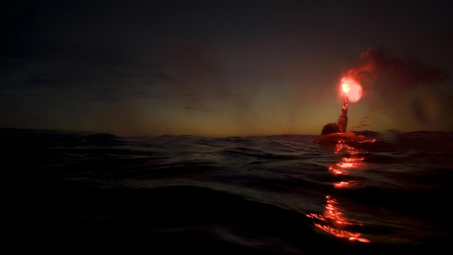 A man in the sea wearing a lifejacket holds up a red flare in the dark sky