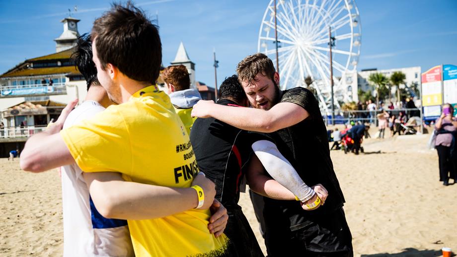 A group of people sharing a congratulatory hug on a beach on a sunny day. They are wearing sports gear and have just finished a tiring activity