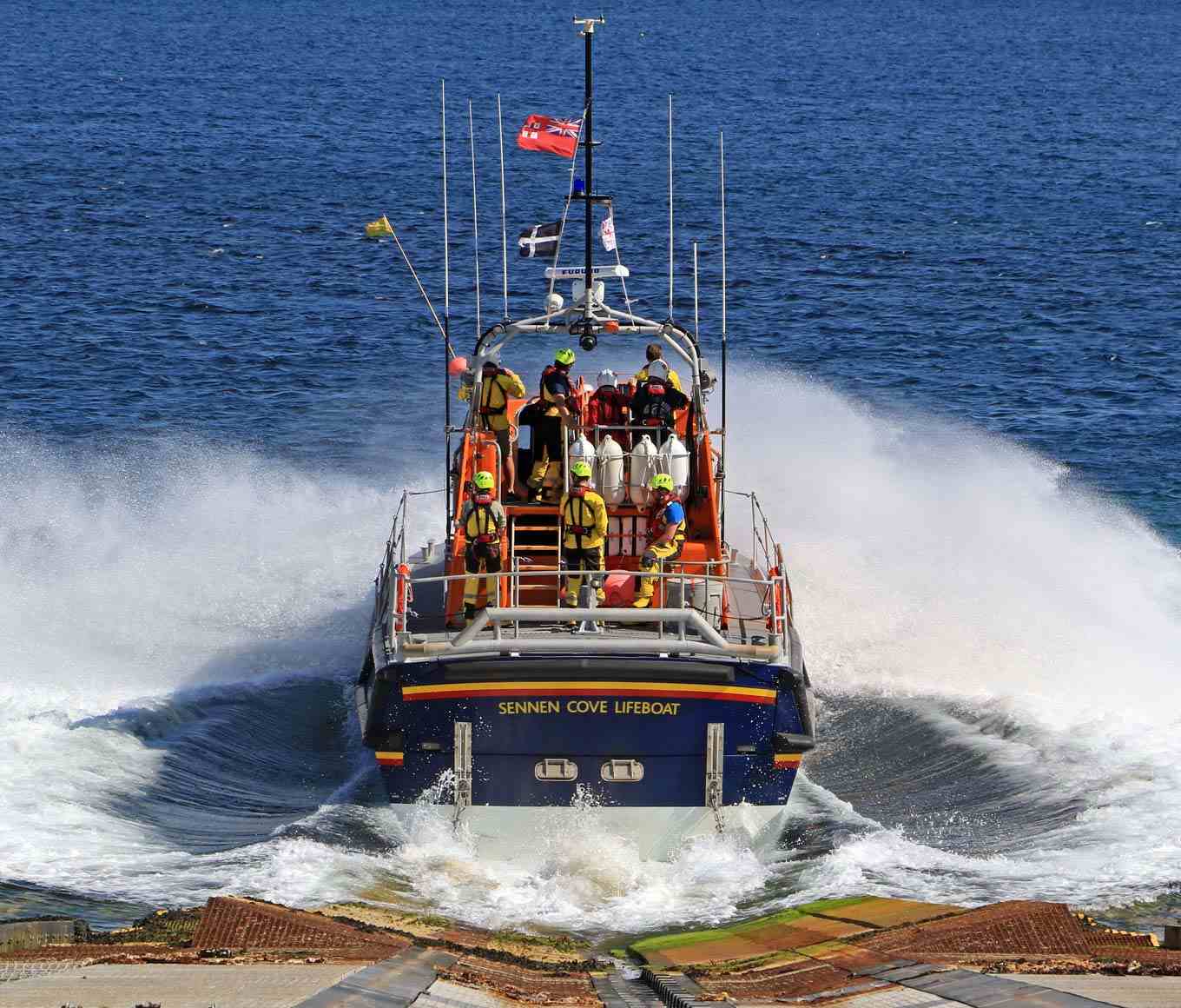 Sennen Cove’s Tamar class lifeboat launches into the waves from the slipway with lifeboat crew in all-weather kit onboard the flying bridge and rear deck