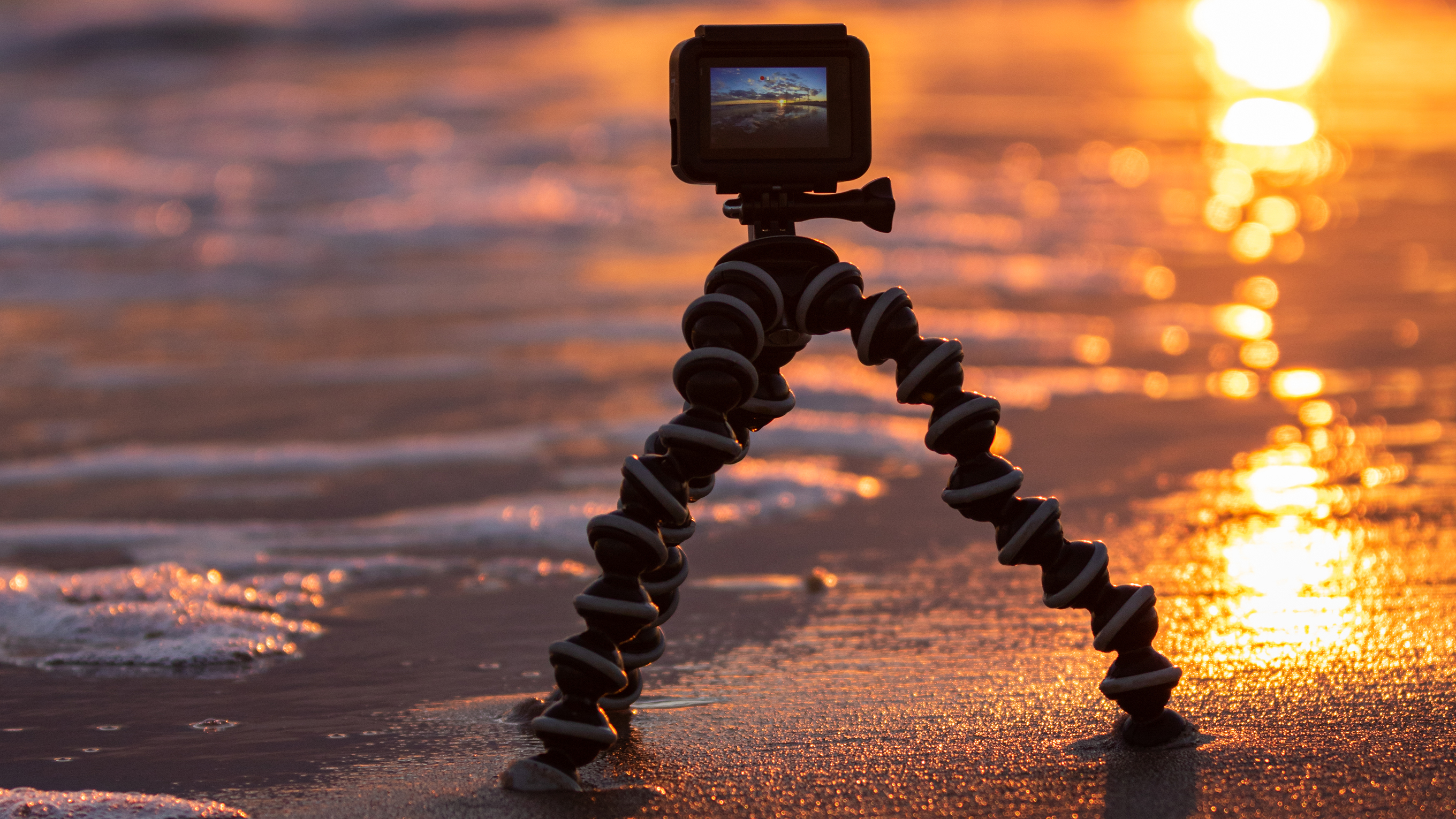 A small camera on a shore-mounted tripod stands on the shoreline. The sun is setting behind it.