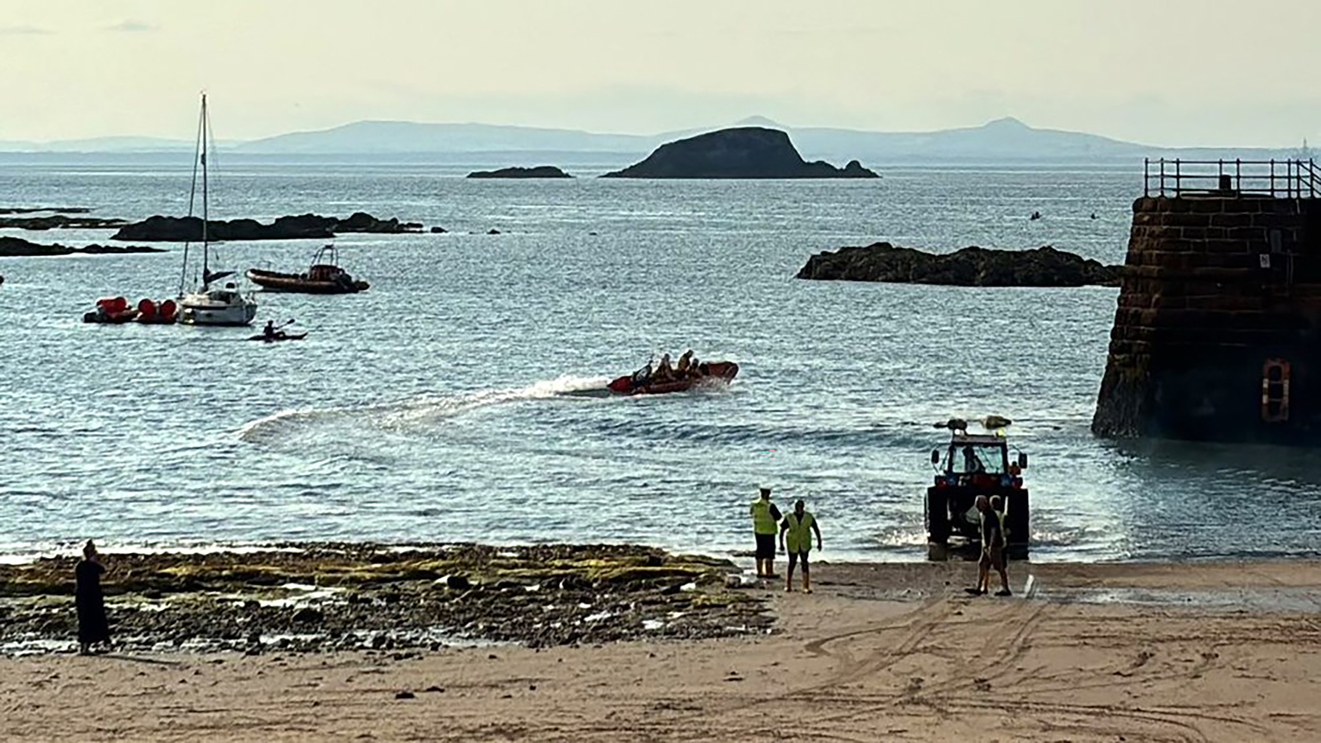 A view across sand and out beyond a harbour wall to the open sea, showing a silhouette of the lifeboat leaving the harbour. The RNLI launching tractor and shore crew are at the water’s edge, seeing them off.