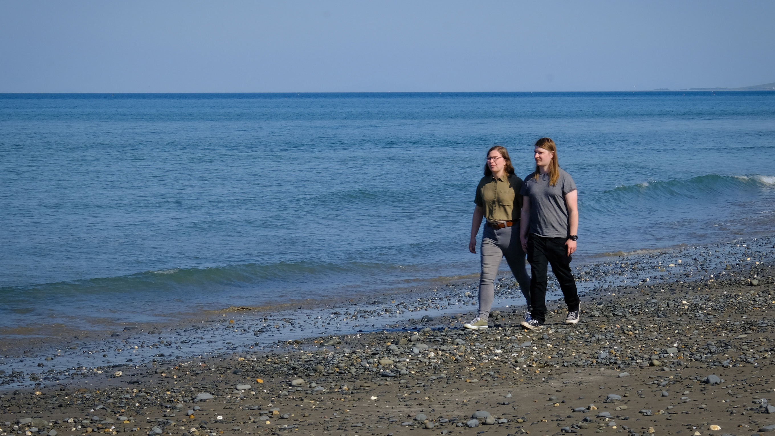 Katie and Will are holding hands and walking on Borth Beach, next to a calm, blue sea.