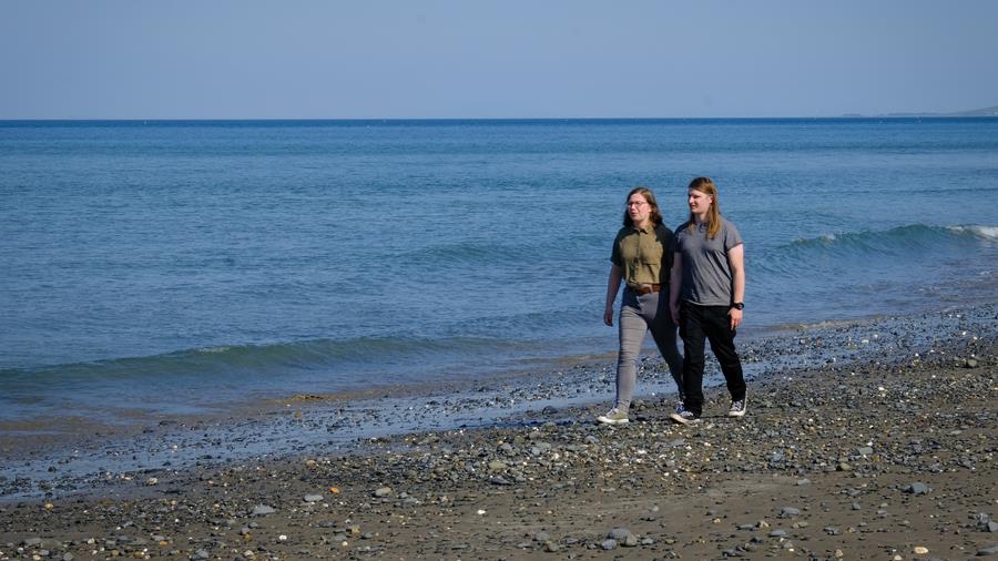 Katie and Will are holding hands and walking on Borth Beach, next to a calm, blue sea.