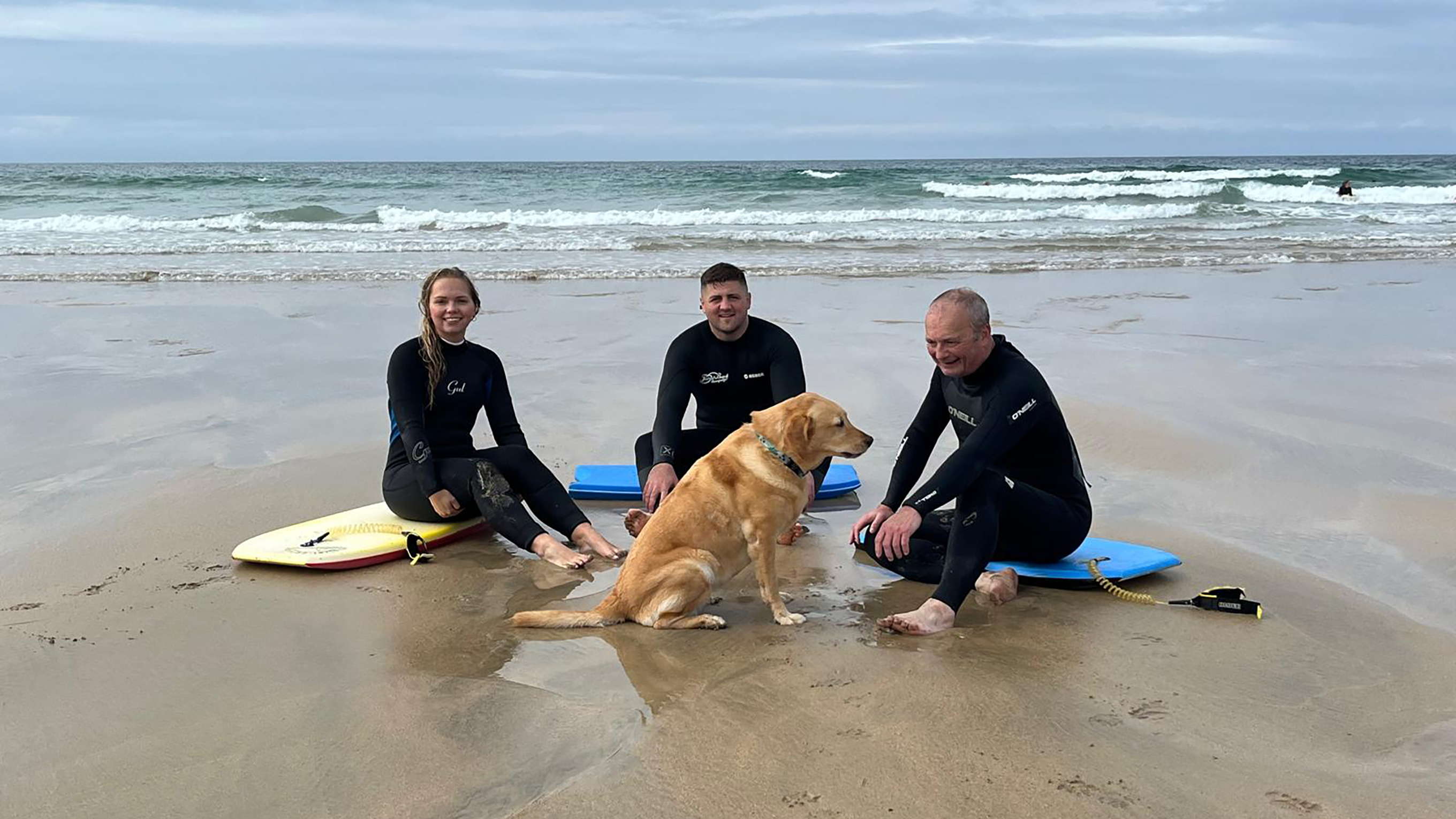 Richard, Charlotte and Ed are sitting on their bodyboards on the sand, wearing wetsuits and still damp from the sea. They’re all smiling and their dog, Flo, sits in the middle of them.
