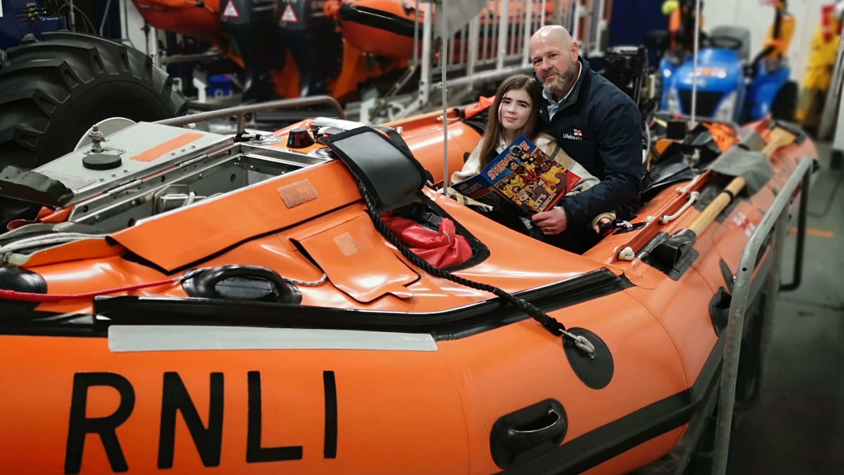 Guy Addington, Margate Crew Member and Regional Water Safety lead, sitting in an inshore lifeboat. His daughter Elodie is sitting in front of him in the lifeboat. Guy is holding a Storm Force magazine in his left hand. Both Guy and Elodie are smiling at the camera. There are other lifeboats and equipment in the background.