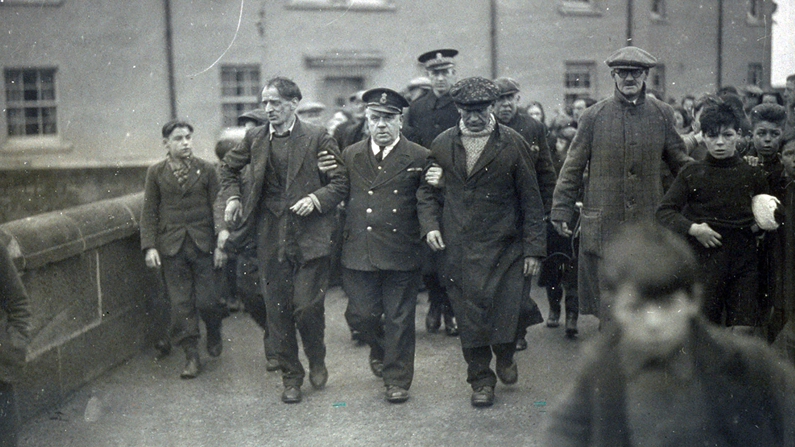 A black and white photograph showing a uniformed coastguard officer holding the arms of two of the crew from the Foremost and helping them walk them down a busy road, which is crowded with people. 