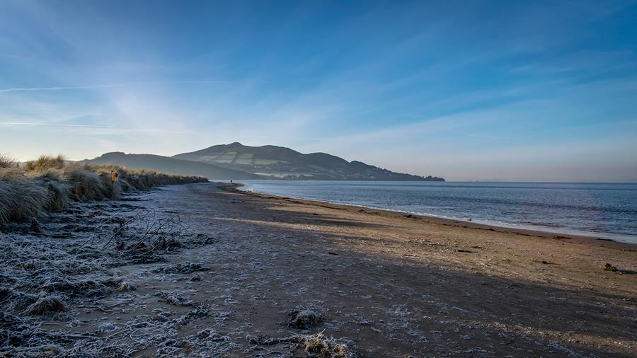 A long sandy beach edged with marshes and covered in frost. Rolling hills with fields and trees can be seen in the distance.
