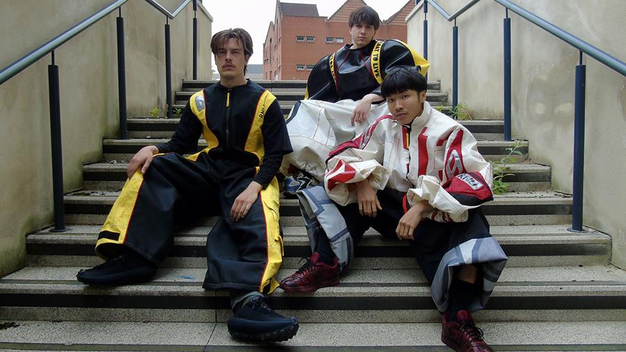 Three young male models dressed in Kaitlyn Joy’s three looks, sitting on some steps in an urban environment.