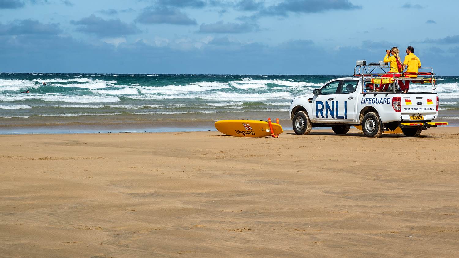 Two lifeguards sit on top of an RNLI vehicle parked on the shoreline of a sandy beach. They are patrolling the choppy waves, and an RNLI rescue board is positioned next to them on the sand.