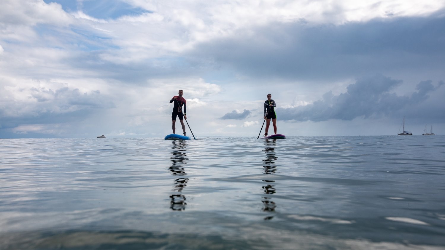 Two paddleboarders out on the water, both wearing wetsuits, paddling towards the camera