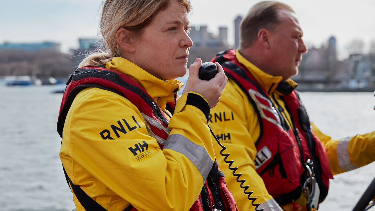 Two lifeboat crew in yellow kit and lifejackets. The female crew member is holding a radio about to speak.