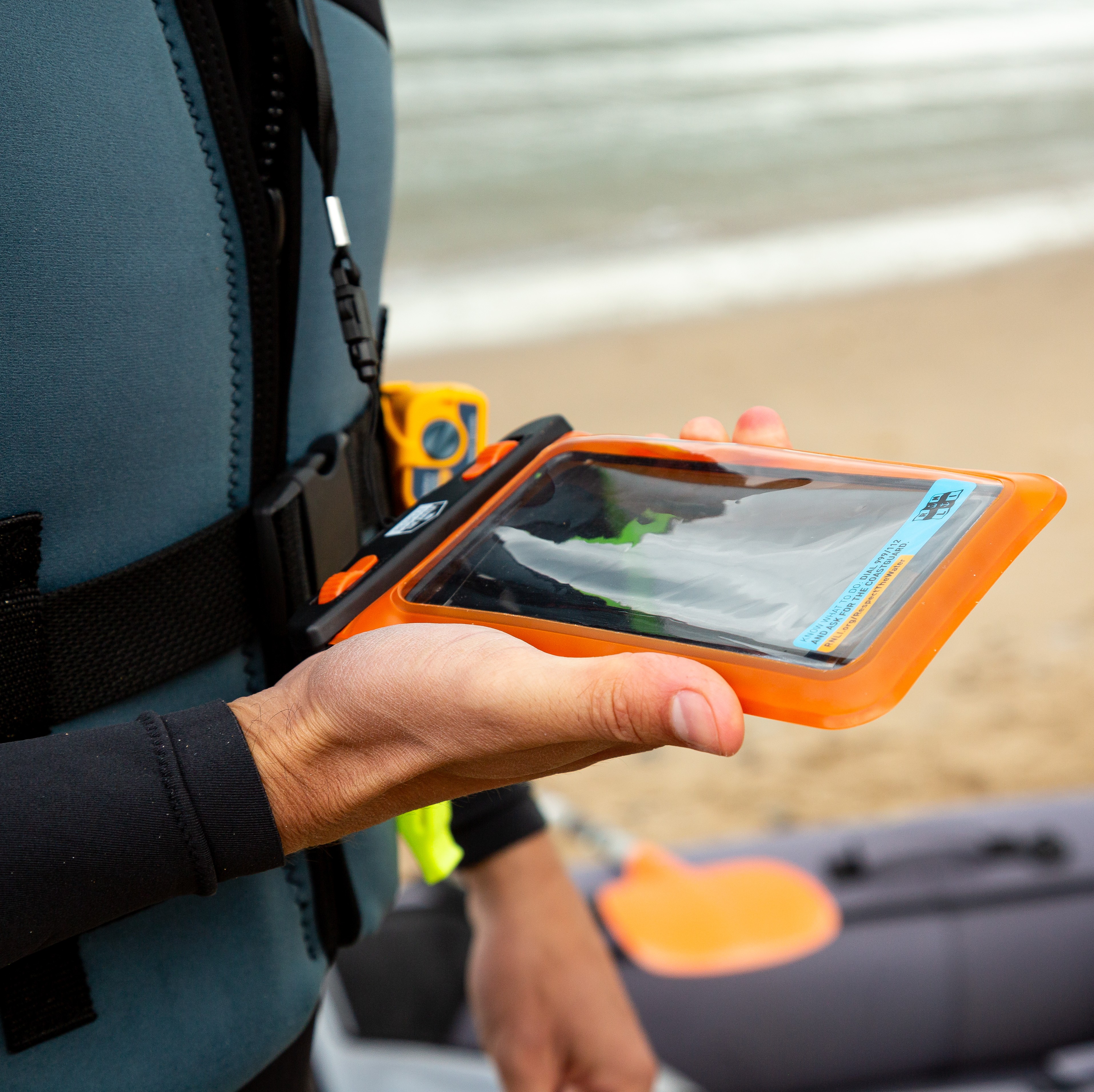 A man is holding a mobile phone in a waterproof pouch on the beach.