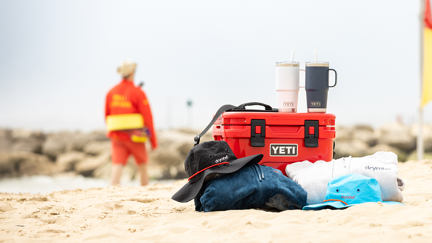 A yeti cooler and two yeti mugs on a lifeguarded beach alongside two dryrobe towel robes and hats