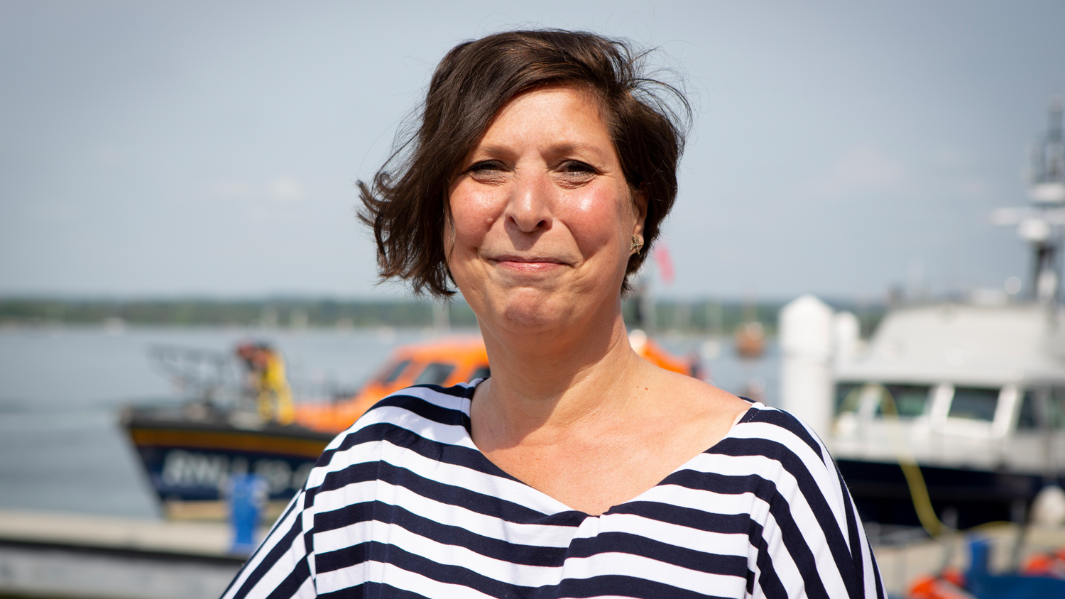 Tina West, who remembered to Float to Live when she got into difficulty in the sea in Cornwall, is smiling at the camera with a harbour behind her on a sunny day. 