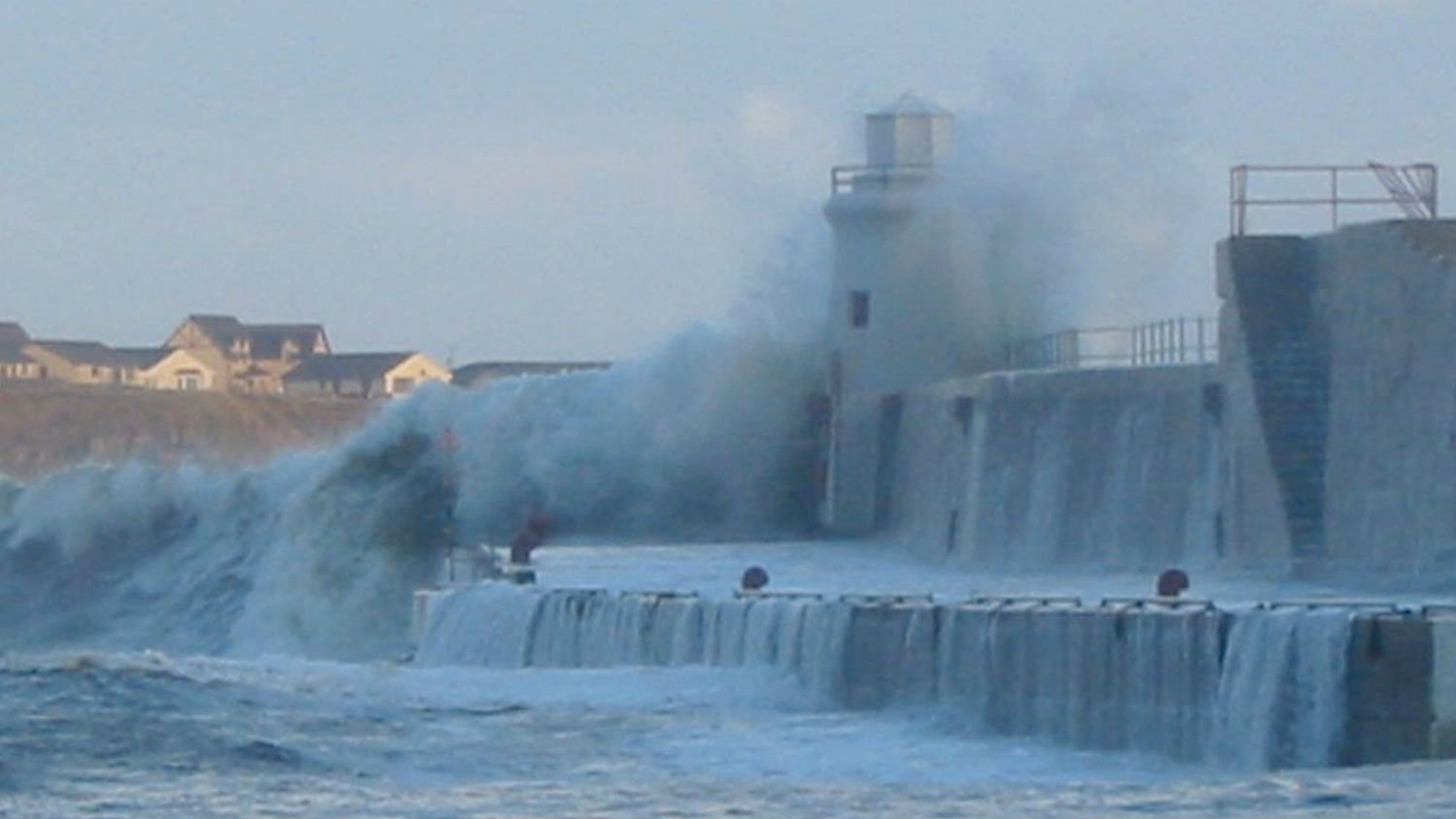Huge wave crashing high over the harbour wall and lighthouse in Wick Bay