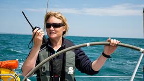 Sailor Pip Hare aboard a yacht, steering the boat and talking into a handheld radio.
