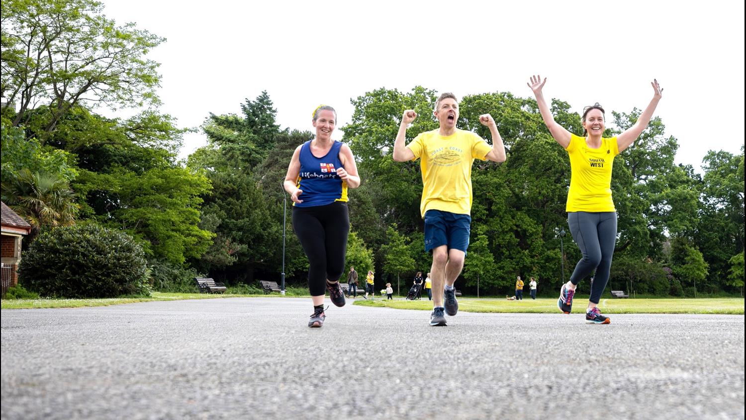 Three people are running along a path in a park for the Mayday Mile. One of them is wearing an RNLI vest, and the other two are wearing yellow T-shirts. They are smiling and cheering. There are trees and grass in the background.