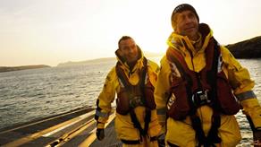 St Davids RNLI Crew Members Roger Smith and Dai John in their all-weather kit walking up the slipway as the sun sets in the distance